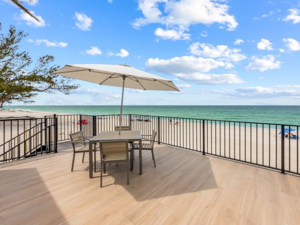 A wooden deck overlooking a beach with a table, chairs, and umbrella under a blue sky with scattered clouds and calm sea.