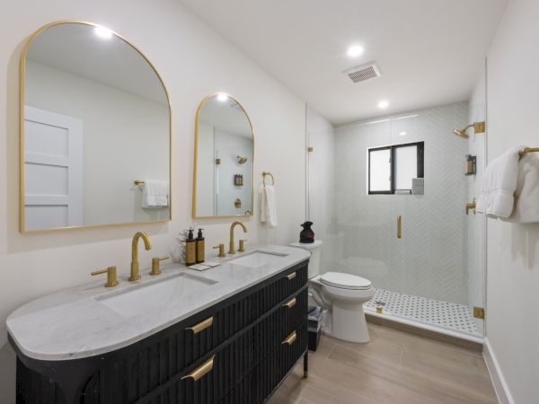 Modern bathroom with dual sinks, gold fixtures, and a glass shower. It features marble countertops and a wood vanity.