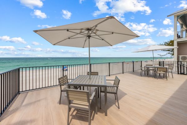 A beachfront deck with tables, chairs, and umbrellas overlooks the ocean under a partly cloudy sky, creating a relaxing outdoor setting.