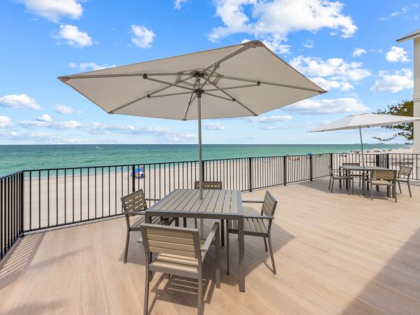 A beachfront deck with tables, chairs, and umbrellas overlooks the ocean under a partly cloudy sky, creating a relaxing outdoor setting.