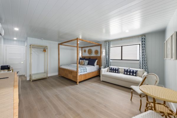 A bright room with a four-poster bed, a sofa, a small table with chairs, and a window with curtains on wooden flooring.