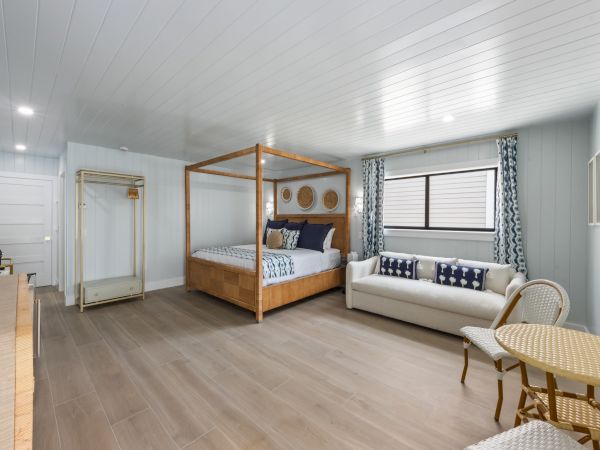A bright room with a four-poster bed, a sofa, a small table with chairs, and a window with curtains on wooden flooring.