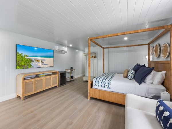 The image shows a well-lit bedroom with a canopy bed, a wall-mounted TV, wooden furniture, and coastal decor themes throughout.