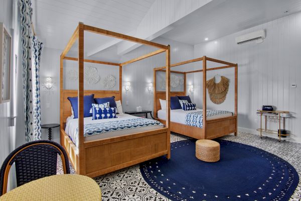 A stylish bedroom with two wooden canopy beds, blue accents, patterned floor, round rug, wicker stool, and wall decor.