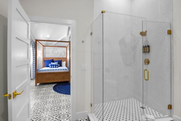 The image shows a bathroom with a glass shower and a glimpse of a bedroom featuring a four-poster bed and patterned flooring.