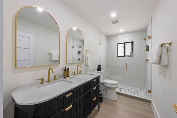 A modern bathroom with double sinks, gold fixtures, and a glass shower enclosure. The design is elegant and minimalist.