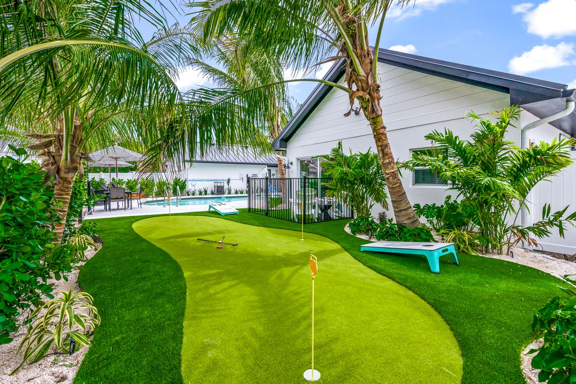 The image shows a tropical backyard with a mini putting green, palm trees, and a view of a pool area by a white house.