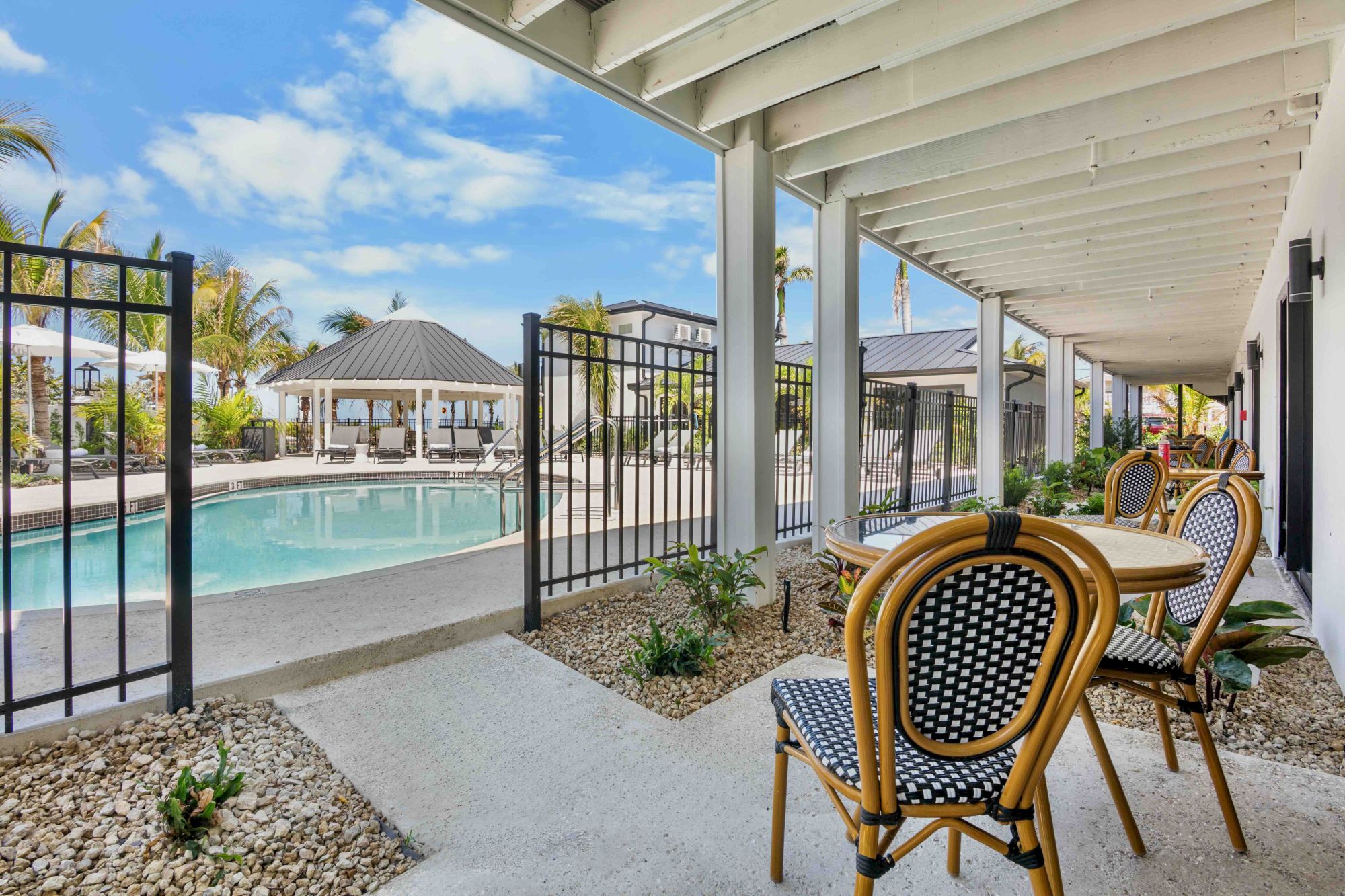 A shaded patio with wicker chairs overlooks a pool, fenced yard, and sunny resort-like outdoor area.