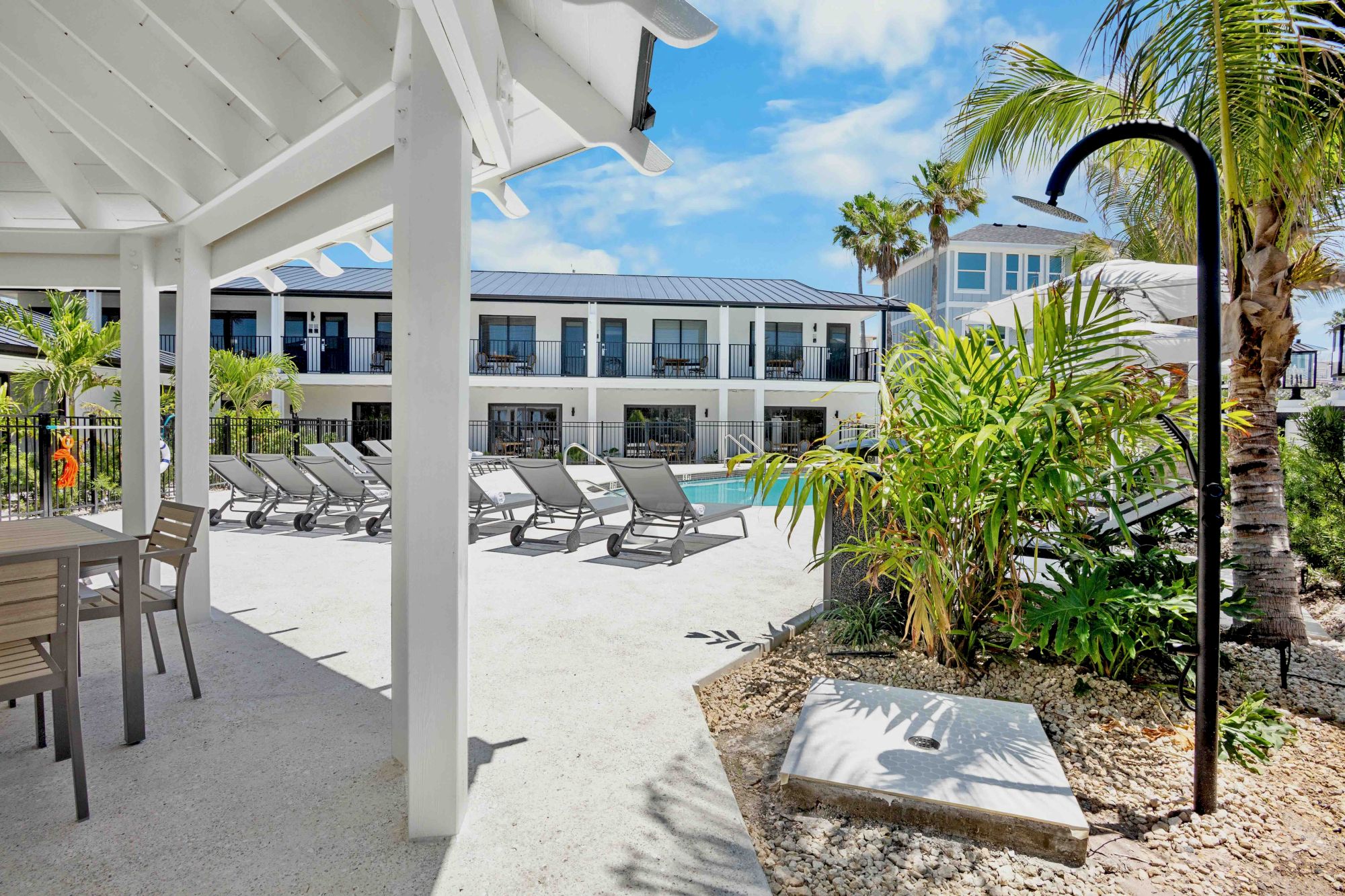 A sunny resort pool area with lounge chairs, palm trees, and a shaded patio, perfect for relaxing in warm weather.