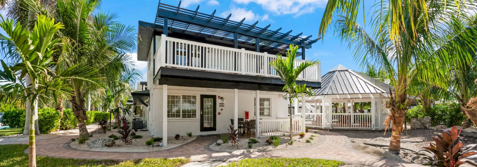 A two-story house with a balcony, surrounded by lush palm trees, and a gazebo to the side, under a clear blue sky.