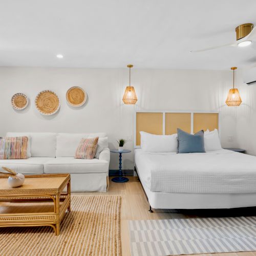 Bright bedroom with a large white bed, sofa, blue curtains, woven wall art, and warm pendant lights, all styled for comfort.