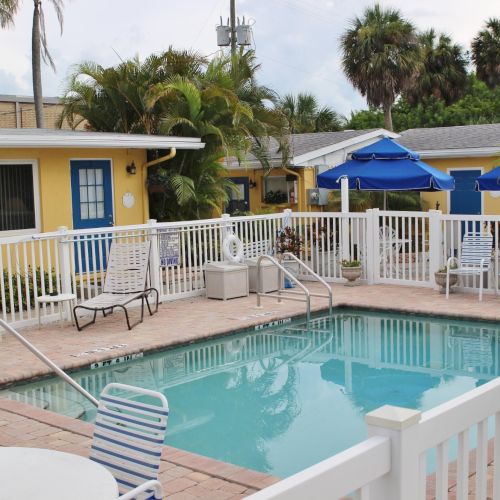 A small pool surrounded by white fencing, lounge chairs, and yellow buildings, with palm trees in the background under a cloudy sky.