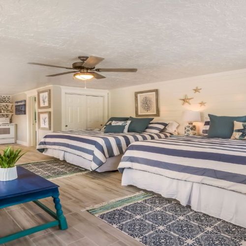 The image shows a spacious room with two striped beds, a kitchenette, and coastal decor. A blue sofa and a coffee table are present.