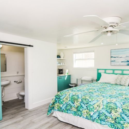 A bedroom with a teal-themed bed, ceiling fan, sliding barn door, and a small adjacent bathroom. Decor includes coastal artwork.