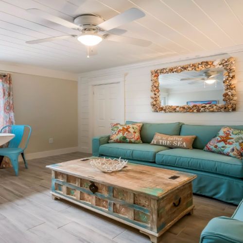 A cozy coastal-themed living room with blue sofas, a rustic coffee table, colorful cushions, a round dining table, and a seashell mirror.