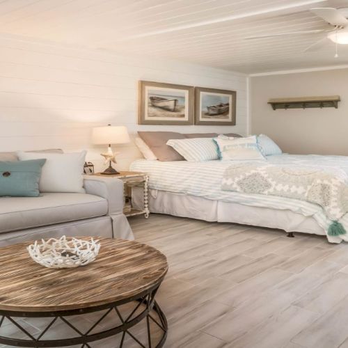 A cozy room with a bed, sofa, wooden table, wall art, and window with curtains. The room has light wood flooring and a ceiling fan.