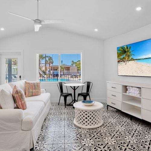 A bright living room with a white sofa, patterned floor tiles, large TV, and window view of a pool area, creating a cozy atmosphere.