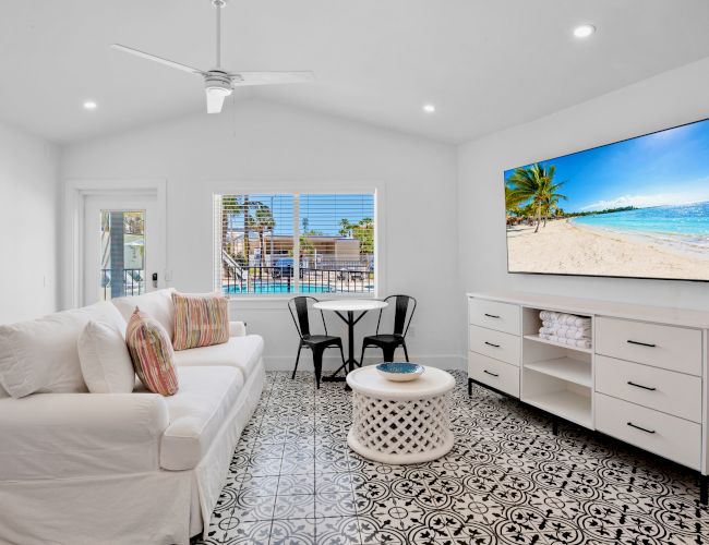 A bright living room with a white sofa, patterned floor tiles, large TV, and window view of a pool area, creating a cozy atmosphere.