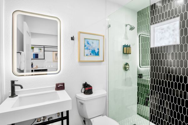A modern bathroom with a lighted mirror, sink, toilet, glass shower, and black hexagonal tiles. Wall art adds a splash of color.