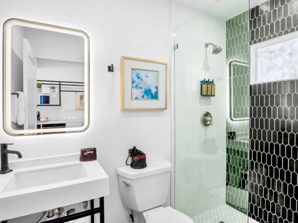 A modern bathroom with a lighted mirror, sink, toilet, glass shower, and black hexagonal tiles. Wall art adds a splash of color.
