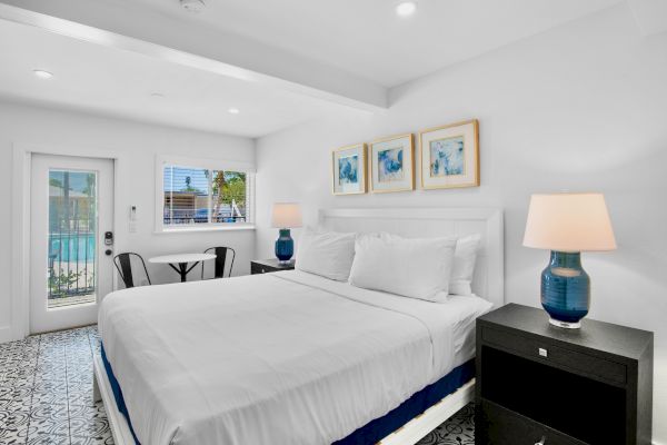 The image shows a bright bedroom with a bed, lamps, window, stylish floor, artwork on the wall, and a small seating area by the door.