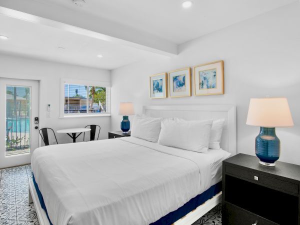 The image shows a bright bedroom with a bed, lamps, window, stylish floor, artwork on the wall, and a small seating area by the door.