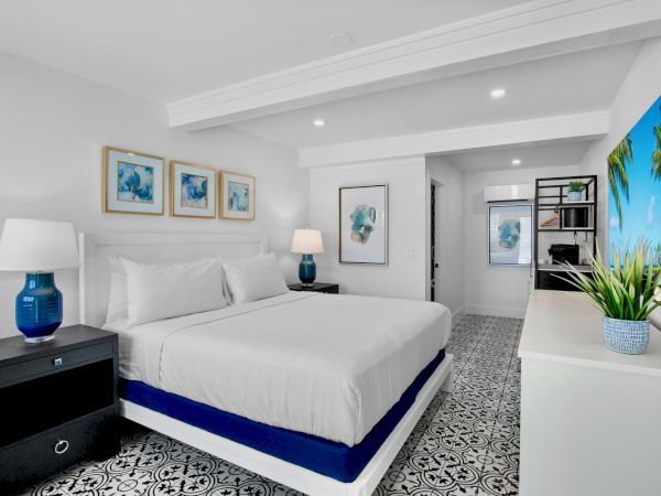 A modern bedroom with a bed, lamps, artwork, and a large TV. The decor includes blue accents and patterned flooring.