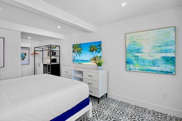 A bright bedroom with patterned floor tiles, a white bed, dresser, and tropical-themed artwork on the walls.