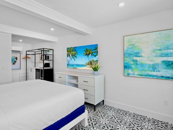 A bright bedroom with patterned floor tiles, a white bed, dresser, and tropical-themed artwork on the walls.