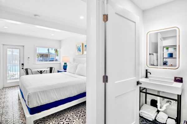 The image shows a bright, modern bedroom with a large bed, adjacent to a compact bathroom featuring a minimalistic vanity and mirror.