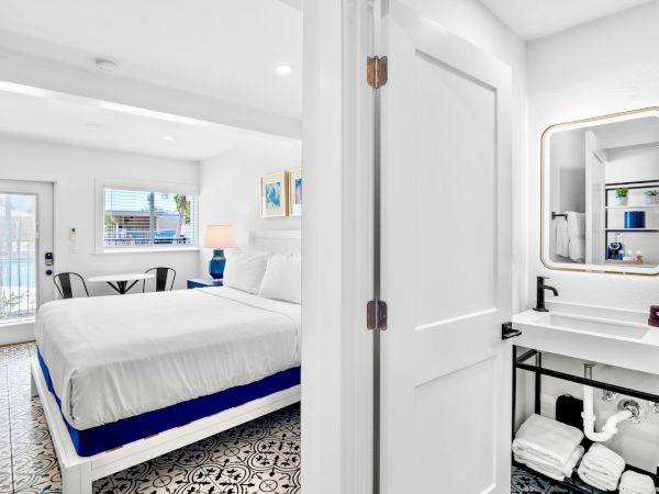 The image shows a bright, modern bedroom with a large bed, adjacent to a compact bathroom featuring a minimalistic vanity and mirror.