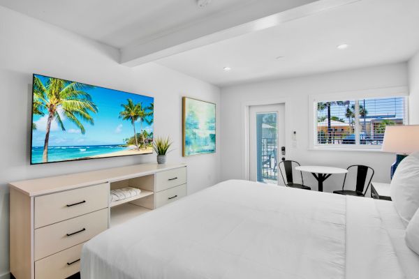 The image shows a bright bedroom with a bed, a dresser, a wall-mounted TV, and a small dining area, featuring a beach scene on screen.