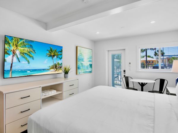 The image shows a bright bedroom with a bed, a dresser, a wall-mounted TV, and a small dining area, featuring a beach scene on screen.