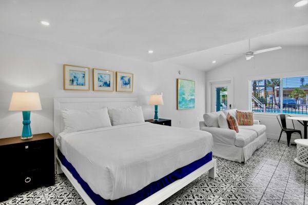 A bright, modern bedroom with a king bed, patterned floor, sofa, wall art, and large window with a view, featuring blue accents.