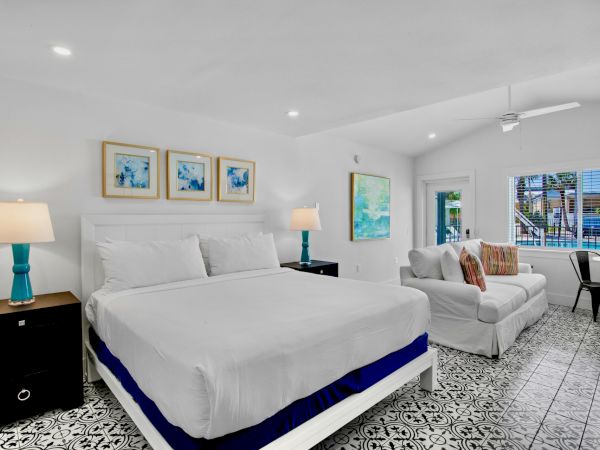 A bright, modern bedroom with a king bed, patterned floor, sofa, wall art, and large window with a view, featuring blue accents.