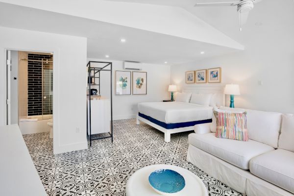 A bright, modern bedroom with patterned floor tiles, a bed, couch, art on the walls, and a bathroom visible.