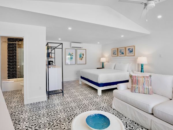 The image shows a modern, stylish bedroom with a patterned floor, a bed, sofa, art on the walls, and a small kitchenette area.