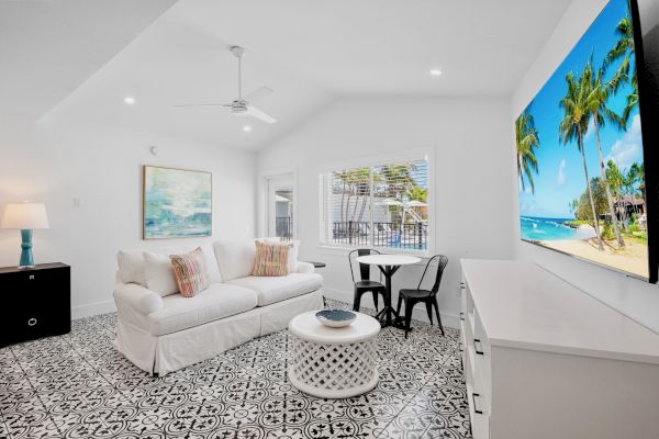 A bright living room features a white couch, stylish patterned floor, and a large wall-mounted TV displaying a beach scene.