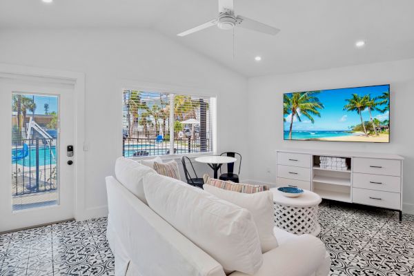 A bright, modern living room with a white sofa, patterned floor, wall-mounted TV, and a view of a pool through the window.