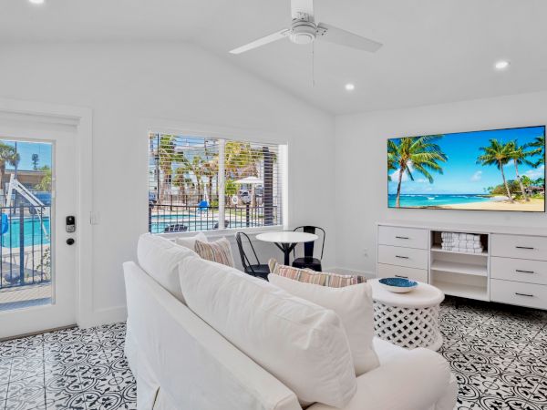 A bright, modern living room with a white sofa, patterned floor, wall-mounted TV, and a view of a pool through the window.