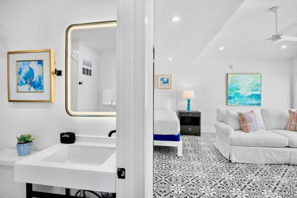 The image shows a modern, bright room with a patterned floor, a sofa, bed, and a bathroom with a sink and mirror.