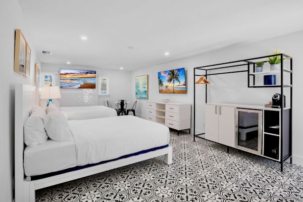 The image shows a modern bedroom with two beds, patterned floor, wall art, storage unit, and a small table and chairs, creating a cozy atmosphere.