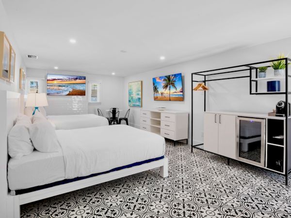The image shows a modern bedroom with two beds, patterned floor, wall art, storage unit, and a small table and chairs, creating a cozy atmosphere.