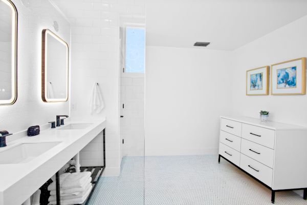 A modern bathroom with dual sinks, lit mirrors, a drawer unit, art on the wall, and a minimalistic white design.
