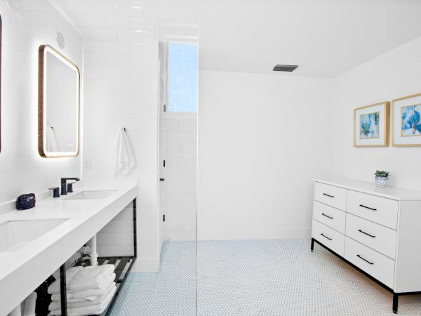 A modern bathroom with dual sinks, lit mirrors, a drawer unit, art on the wall, and a minimalistic white design.
