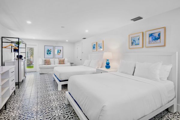 A spacious, bright bedroom with two beds, decorative blue art, a couch, tile flooring, and a lamp on the nightstand, exuding coziness.