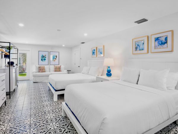 A spacious, bright bedroom with two beds, decorative blue art, a couch, tile flooring, and a lamp on the nightstand, exuding coziness.