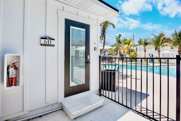 A white building with a door labeled "611," a fire extinguisher, and a gated pool area with palm trees under a blue sky.
