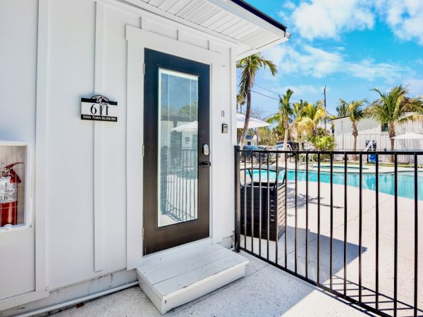 A white building with a door labeled "611," a fire extinguisher, and a gated pool area with palm trees under a blue sky.