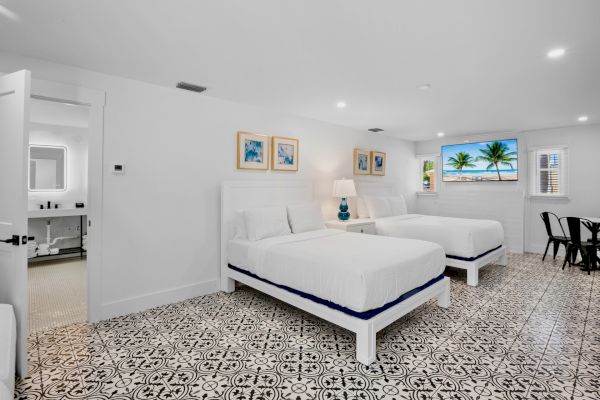 A bright bedroom with two white beds, decorative floor tiles, paintings, a lamp, and a window with a scenic view completes the space.